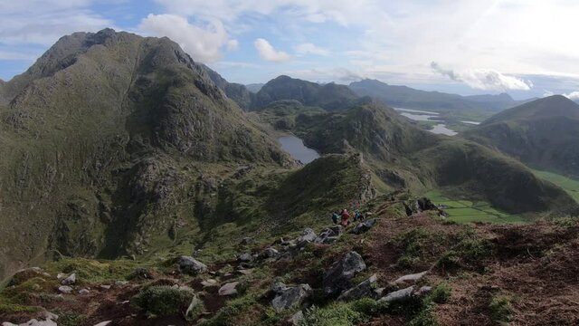 A group of hikers are walking down the trail from the top of mountain Mannen at Haukland beach in Lofoten, Norway. Fjords, valleys and mountains in the epic and majestic background.