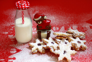 Snowflakes shaped Christmas cookies on a red background with powdered sugar silhouettes of houses under the cookies and a milk bottle with straw in it, and a chocolate bear shape dessert