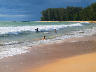 Tropical resort, swimming people on the beach, forest in a distance on a horizon. Dark blue cloudy sky and tourists resting on the coast. Yellow sand, azure sea, waves. Panoramic view of Phuket beach.