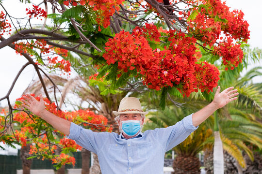 A senior man white hair and cap with open arms and face mask due to coronavirus enjoys outdoor under flowering plants in the park - concept of new normality after lockdown for Covid-19 man - Powered by Adobe