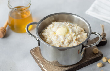 close-up of boiled oatmeal porridge in a pan, hazelnuts and a jar of honey on a gray table. delicious homemade breakfast. horizontal image.