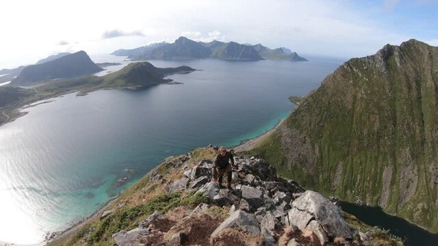 A young man with a camouflage jacket is hiking up the last steps to the peak and top of Mannen mountain. Beautiful view overlooking Haukland beach in Lofoten, Norway.