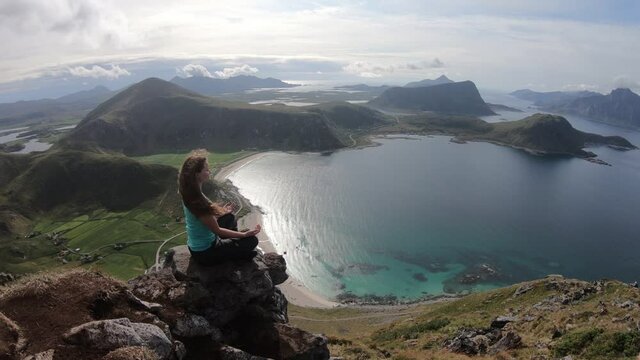 A young hiker woman is sitting down to meditate on top of mountain Mannen, overlooking Haukland beach. Feeling of peace, tranquillity and harmony as the wind blows in her hair.