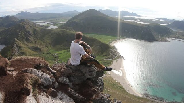 A young tattooed man is sitting by himself, looking at the beautiful view of Haukland beach below. Peaceful and quiet time on top of mountain Mannen in Lofoten, Norway.