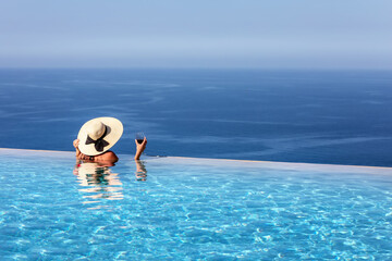 Eine attraktive Frau mit Hut und Weinglas in der Hand genießt die Aussicht vom infinity Pool auf...