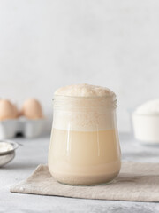 Homemade sourdough yeast starter bubbling in glass jar, sieve, eggs, bowl with flour. food ingredients. vertical image. Gray background. copy space. selective focus