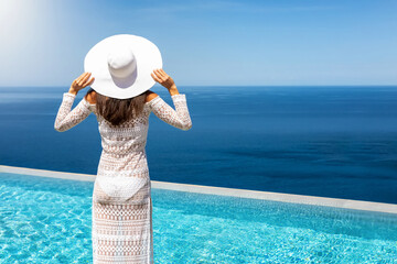 Fototapeta na wymiar Eine Frau mit weißem Hut und Sommerkleid steht am Pool und schaut auf das blaue Meer