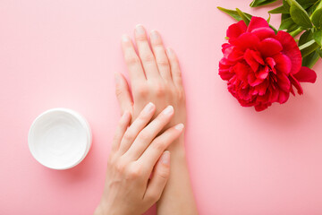 Young woman hands and opened white cream jar on light pink table background. Pastel color. Beautiful red peony flower. Care about clean and soft body skin. Daily beauty product. Closeup. Top view.