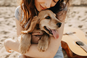 Young woman sitting on the sand on a beach, hugging her little dog, cocker spaniel breed puppy.
