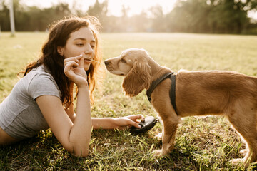 Young woman training and feeding her little dog, cocker spaniel breed puppy, outdoors, in a park.