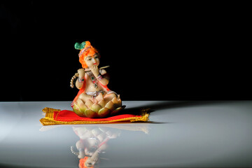 An isolated sculpture of Indian god lord Krishna playing flute sitting on a mat. On a reflective white table with a dark background © Suman