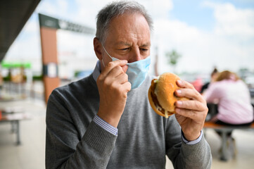Mature man trying to eat a hamburger wearing a mask, funny coronavirus concept