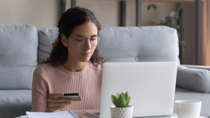Focused woman wearing glasses paying online by plastic credit card, looking at laptop screen, serious girl shopping buying online, customer making internet payment, sitting at table in living room