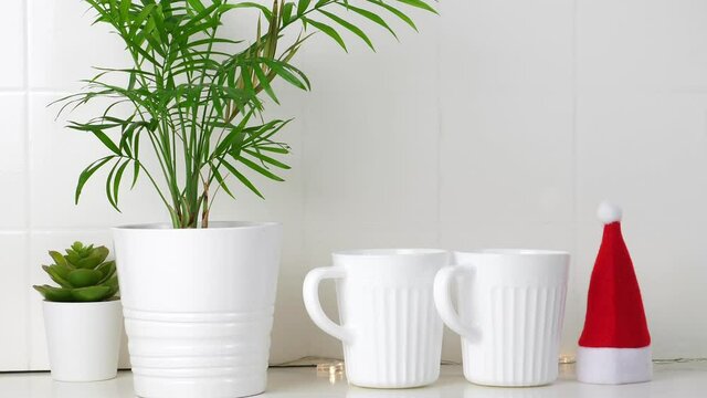 Santa Claus hat, Tea cups, green palm plant and succulent on white kitchen table. Closeup. Nobody