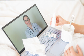 A woman lies in bed and holds a spray for sore throat. A friendly doctor talks to a sick patient by video link. Therapist on a laptop screen. Online medical consultation during the quarantine.