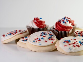 Pile of sugar cookies with white frosting and red white and blue sprinkles with mini chocolate cupcakes frosted in red and white with star sprinkles on a white counter for 4th of July.