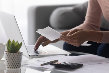 Close up woman holding receipt, using laptop, calculating bills, money, sitting at table on couch,...