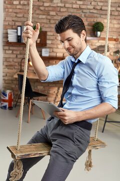 Businessman sitting on swing, using tablet in office. 