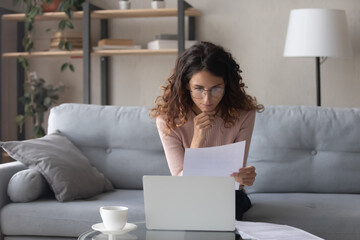 Thoughtful woman wearing glasses reading document, using laptop, sitting on couch in living room,...