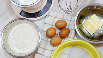 Baking ingredients of brown eggs, white flour, butter, flour sifter, kitchen scale and a metal whisk, placed on a table top.
