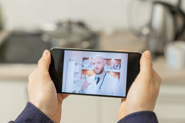 Fototapeta na wymiar A serious bearded doctor communicates with a patient via a video call on a smartphone. Online medicine concept. Woman in the kitchen takes therapist remote consultation on mobile.