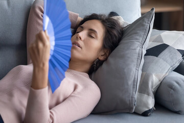 Exhausted overheated woman waving blue paper fan close up, lying on couch, relaxing on soft...