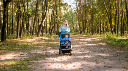 Happy smiling young mother with baby pram walking on pathway at autumn forest