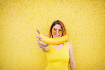 Beautiful red-haired woman in sunglasses holds a banana and fools around. Attractive European girl in a dress posing on a yellow background charmingly smiles.