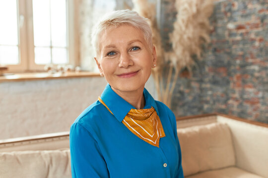 Indoor Image Of Beautiful Middle Aged Woman Wearing Blue Shirt And Neckerchief Relaxing At Home, Sitting On Comfortable Sofa, Smiling At Camera, Having Happy Relaxed Look. People And Lifestyle