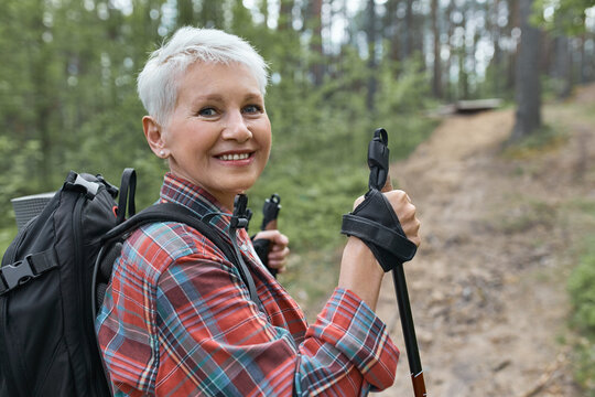 Active lifestyle, aging, travel and hiking concept. Outdoor image of beautiful energetic mature woman with backpack using poles, enjoying nordic walking in forest, looking at camera with happy smile