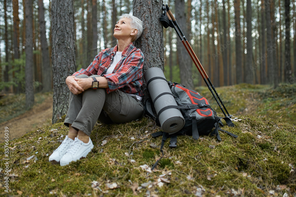 Wall mural beautiful mature woman in sneakers and activewear sitting on grass under pine having rest during nor