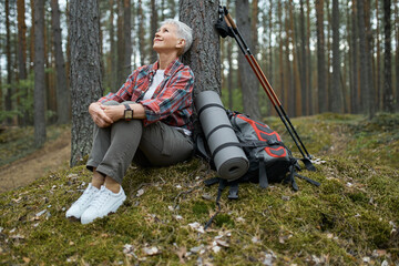 Beautiful mature woman in sneakers and activewear sitting on grass under pine having rest during nordic walking with sticks and backpack, looking up with relaxed carefree smile, breathing fresh air