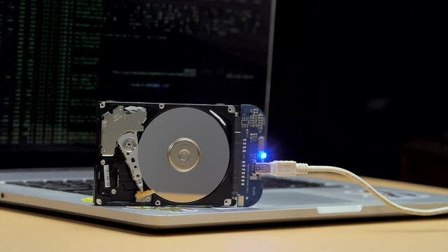 Hard drive stands on the laptop keyboard, flashes and disk spins. Work of hacked hard drive inside. Hacker breaks sensitive data from hard drive. Lines of code and data flash in monitor on background.