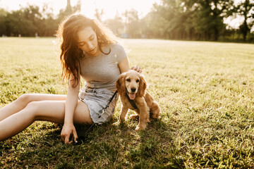 Girl sitting on green grass with her little dog, cocker spaniel breed puppy, outdoors, in a park.