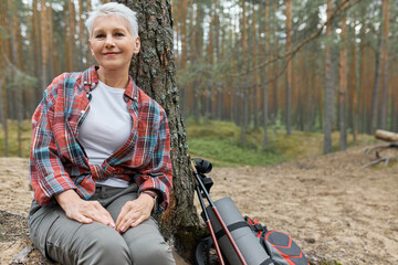 Attractive Caucasian mature woman in activewear relaxing under tree with backpack next to her, smiling, catching breathe while hiking in woods, enjoying peace and tranquility of wild nature
