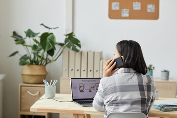 Back view portrait of modern young woman speaking by smartphone and using laptop while sitting by wooden table and working at home office, copy space