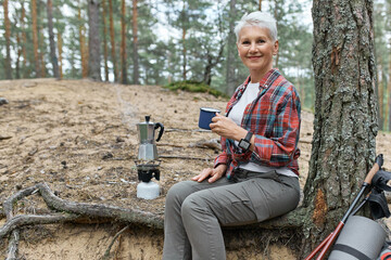 Outdoor summer image of cheerful middle aged woman in activewear relaxing under tree with camping...