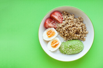 Bowl of oatmeal porridge with avocado, tomato, boiled egg and sesame on green background. Oatmeal with vegetables. Hot and healthy breakfast and diet food. Balanced nutrition concept