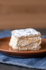 Cremșnit sitting on top of a plate. Is made out of filo pastry of usually three layers with cream on top and crumble sprinkled on it.
