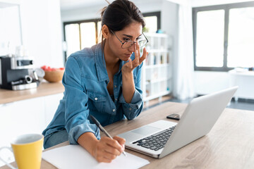 Pretty young woman writing down notes and working on a laptop in the kitchen at home.