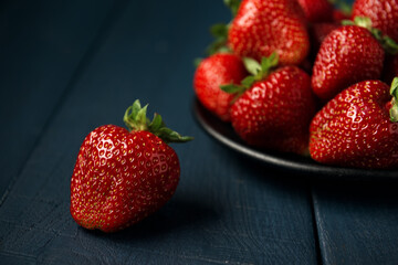 Ripe red strawberry and group of strawberries in a black plate on a dark blue wooden background. Dessert from fresh berries on the table.