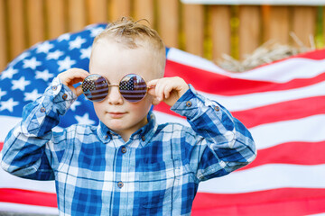 Cute blond boy in stylish glasses stands in the yard on the background of the American flag