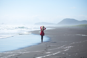 Beautiful young blonde girl in red dress is walking and posing on the beach of Pacific Ocean with black volcanic sand.