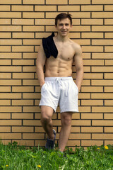 A bare-chested athletic young man in white shorts is standing by an orange brick wall.