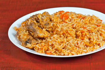 Pilaf with meat and carrots in a white plate. Rice on red plywood.