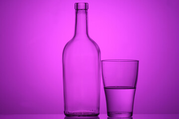 Photo of a bottle and half full glass with backlighting on a purple background. stacked objects