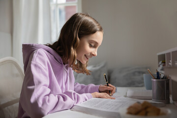 Smiling smart teenage schoolgirl sit at desk at home do prepare homework with textbook alone, happy teenager girl pupil study learn in bedroom handwrite in notebook, distant school education concept