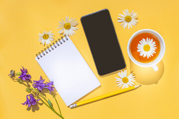 Notebook on a spiral with an empty sheet, a smartphone, a yellow pencil, a cup of tea and chamomile flowers on a bright yellow background. Copyspace, minimalism, top view. Summer cute funny flat lay.