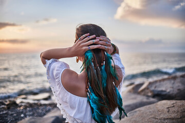 Hippie woman with blue feathers in hair on the seashore at sunset. Bohemian vibes and boho chic...