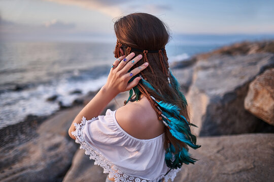 Hippie indie woman with blue feathers in hair on the seashore at sunset. Boho vibes and bohemian fashion